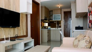 A modern living room with a flat-screen TV, a wooden entertainment unit, a white sofa with patterned cushions, and a kitchen area in the background. - Brand New And Modern 1Br Signature Park Grande Apartment (Jakarta)