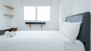 A neatly made bed with white linens, a dark headboard, and two white pillows. There is a window with a white blind, and a small wooden shelf unit to the left. - Modern 1Br Apartment At Signature Park Grande (Jakarta)