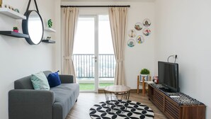 A modern living room with a grey sofa, a round coffee table, a TV on a wooden stand, and a balcony with curtains. - Modern 1Br Apartment At Signature Park Grande (Jakarta)