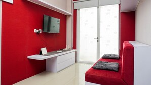 Une chambre à coucher aux couleurs rouge et blanc, avec un téléviseur fixé au mur, un bureau avec une chaise, un lit avec des oreillers et une porte-fenêtre coulissante donnant sur un balcon.
