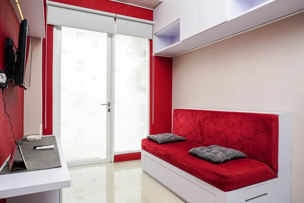 A room with a red sofa bed, a white desk, and a large window with a view of trees.
