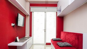 Une pièce moderne avec un mur d’accent rouge, un bureau blanc avec un ordinateur, un canapé rouge agrémenté de coussins et une porte-fenêtre coulissante donnant sur un espace lumineux.