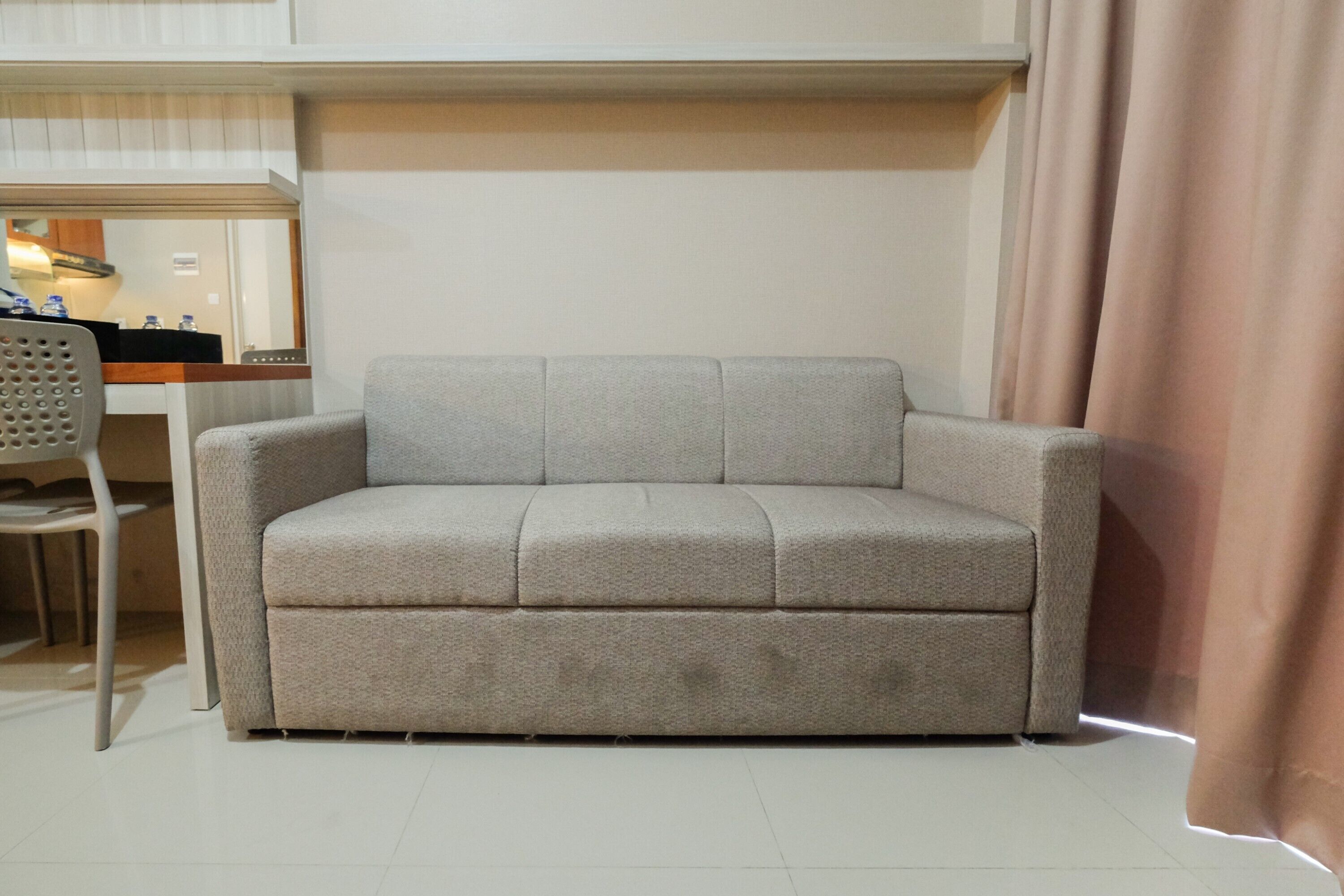 Un canapé gris clair dans une pièce dont le sol est recouvert de carreaux blancs et où se trouve un bureau en bois avec une chaise.