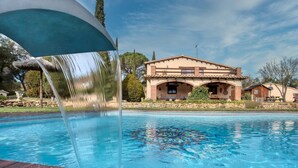 A swimming pool with a waterfall feature, a house with a tiled roof, and a stone wall in the background.