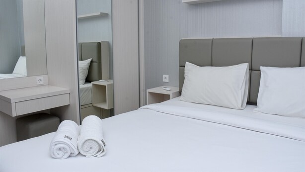 Una cama bien hecha con sábanas y almohadas blancas, una mesita de noche con un control remoto, una pequeña mesita auxiliar y un espejo sobre la cama.