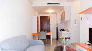 A modern hotel room with a grey sofa, a dining table with a vase of flowers, a kitchen area, and a TV on a stand.