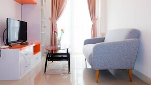 A modern living room with a flat-screen TV on a white cabinet, a grey armchair, a small round table with a vase, and a window with curtains.