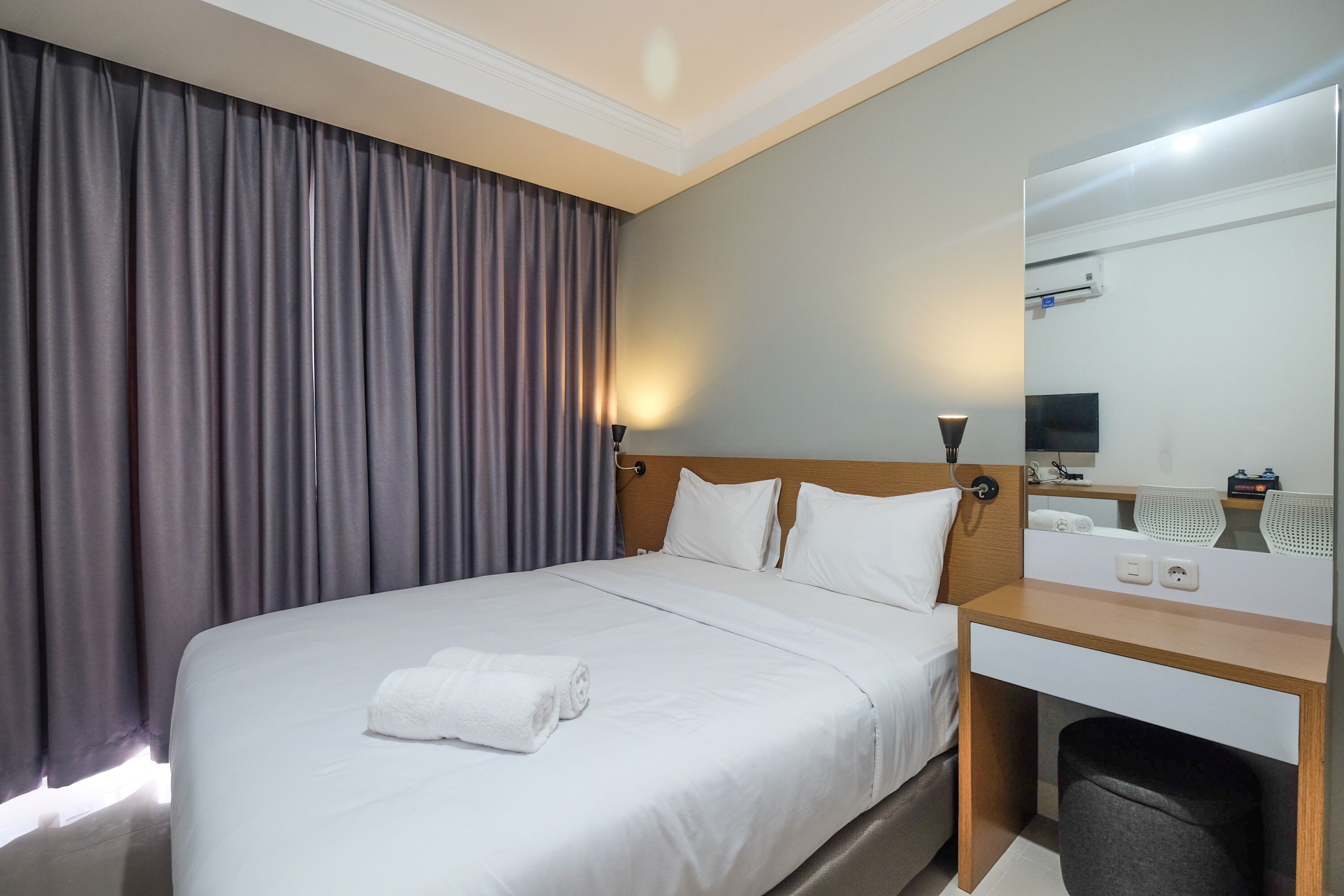 A hotel room with a large bed, a wooden desk, a mirror, and a window with curtains.