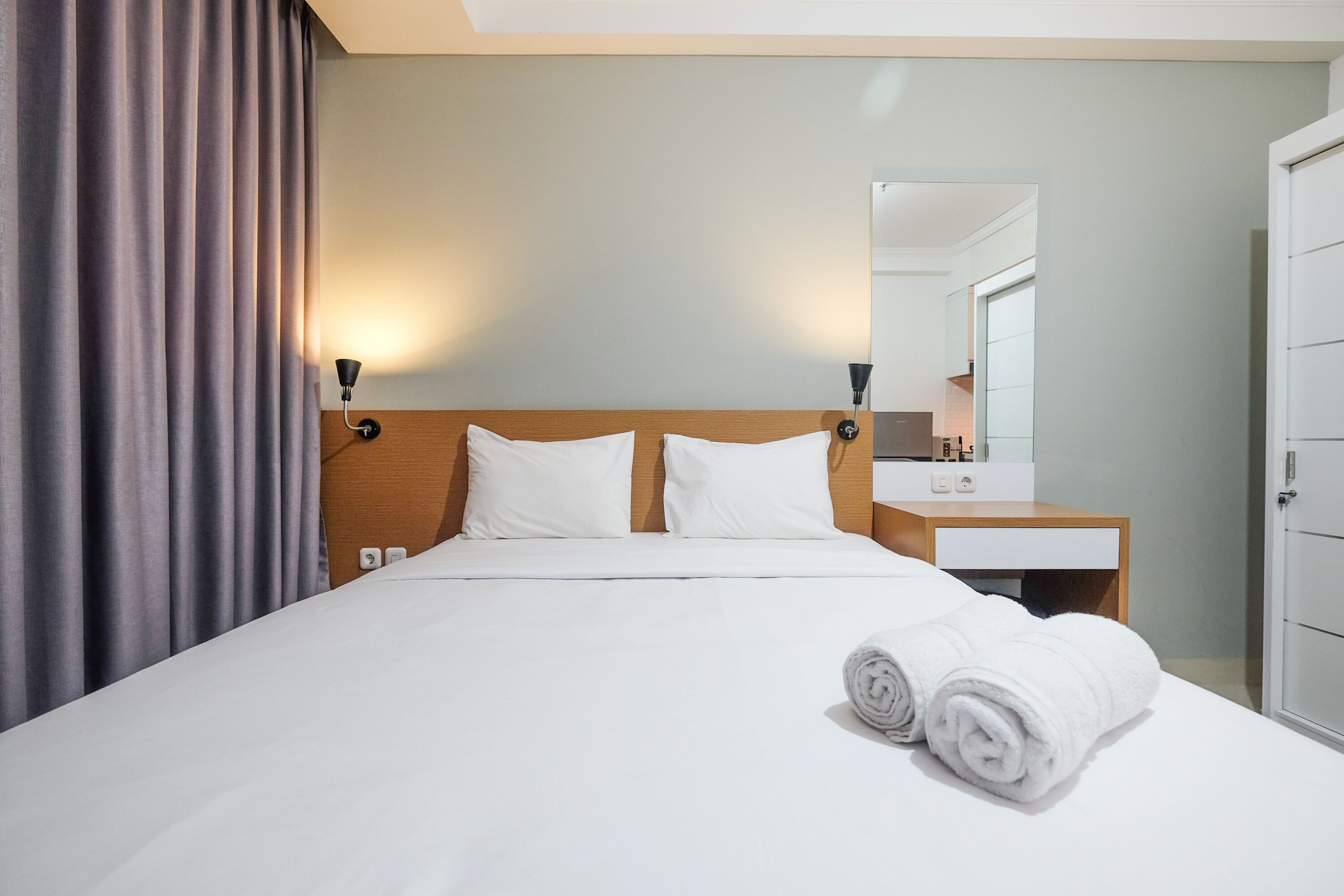 A hotel room with a neatly made bed, a wooden headboard, a bedside table with a lamp, a mirror, and a wardrobe.