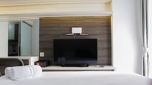 Habitación de hotel con un televisor de pantalla plana grande montado en una pared de madera, una mesita de noche con un control remoto, una toalla blanca enrollada y un espejo que refleja la habitación.