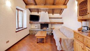 Ein gemütliches Zimmer mit Kamin aus Stein, Holzbalken an der Decke, einem Fernseher und einer Couch.