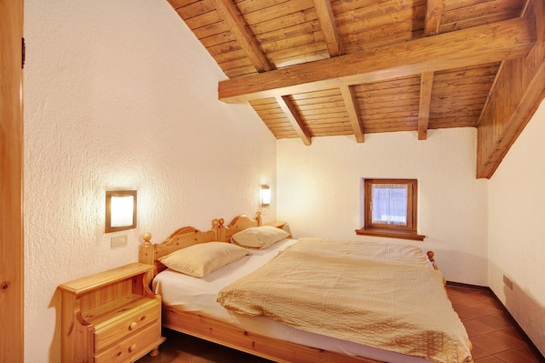 A bedroom with a wooden bed, nightstand, and dresser, wooden ceiling beams, and a window with curtains.