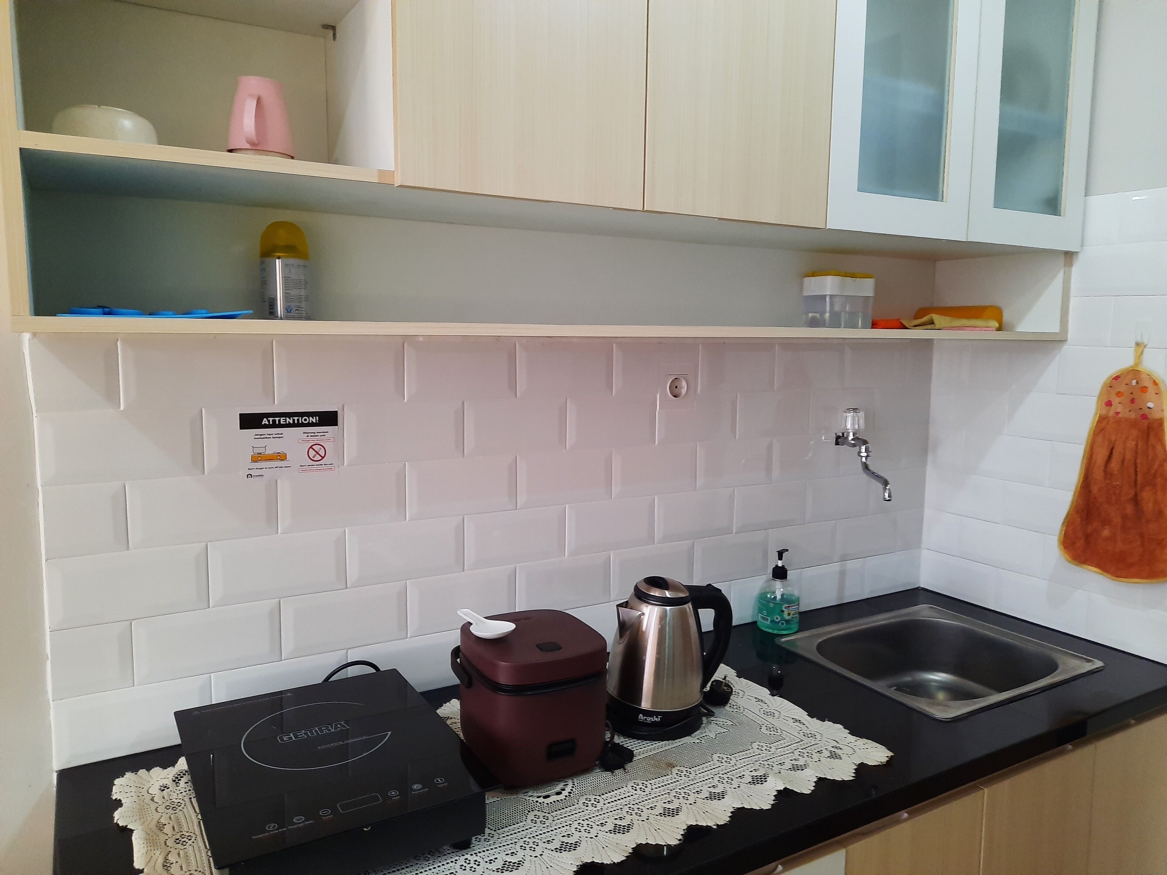 A kitchen with a black countertop, a stainless steel sink, a kettle, a coffee maker, and a microwave. There is a white tiled backsplash and a sign on the wall.