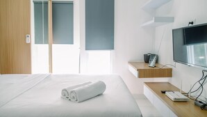 A modern hotel room with a large bed, a TV, a desk with a computer, and a window with blinds. - Stunning And Comfy Studio Amazana Serpong Apartment (South Tangerang)