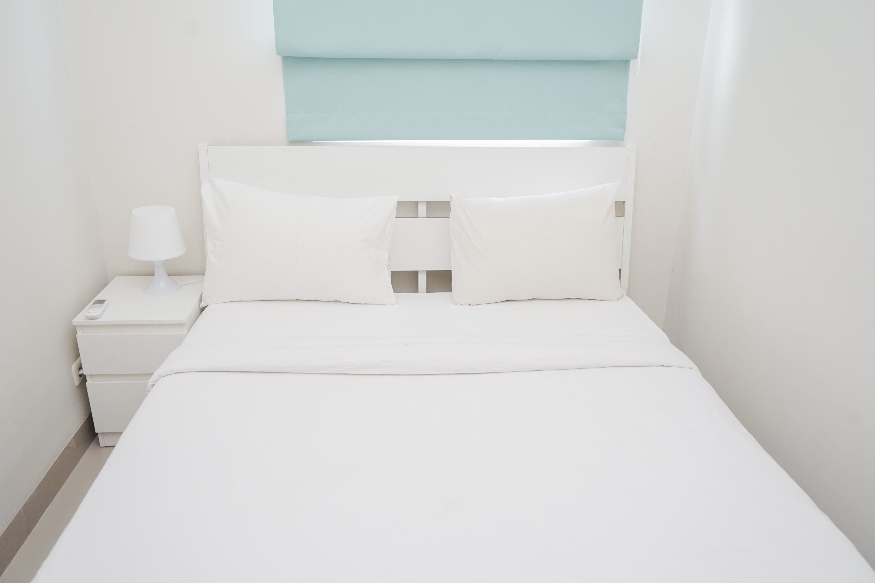 A small, white, single-bed room with a nightstand, a lamp, and a window with a light blue blind.