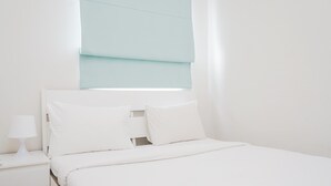 A neatly made bed with white bedding and pillows, a bedside table with a lamp, and a window with a light blue blind. - Cozy And Spacious 1Br Apartment At Parkland Avenue (South Tangerang)