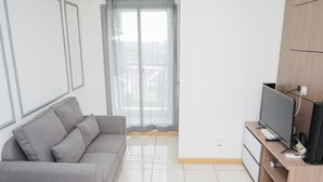 Una sala moderna con un sofá gris, un televisor de pantalla plana sobre un soporte de madera y un ventanal corredizo que da a un balcón.