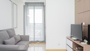 Una sala de estar moderna con un sofá gris, un televisor de pantalla plana sobre un soporte de madera y un balcón con cortinas translúcidas.