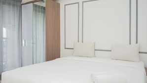 Una cama bien tendida con sábanas y almohadones blancos, un armario de madera y una puerta de cristal que da a un baño.
