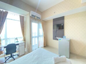 A hotel room with a bed, desk, chair, air conditioning unit, television, and a water dispenser. - Modern Studio Room At Taman Melati Jatinangor Apartment (Hegarmanah)