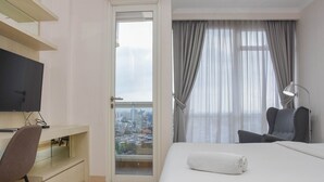 A hotel room with a bed, a desk, a chair, a TV, and a view of the city through a large window. - Comfortable And Warm Studio Room At Menteng Park Apartment (Jakarta)