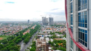 A cityscape with a prominent building featuring a red facade and large windows, surrounded by other high-rise buildings and a mix of residential and commercial structures. - Warm Studio Apartment At Tamansari Papilio (Surabaya)
