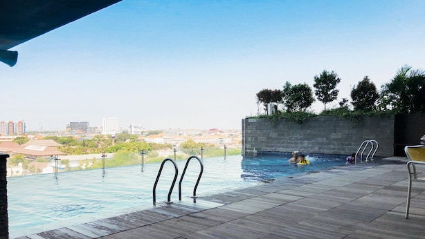An outdoor infinity pool with a city view, a wooden deck, and a stone wall. - Warm Studio Apartment At Tamansari Papilio (Surabaya)