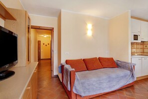 A modern living room with a flat-screen TV, a sofa with a blanket, a kitchen area with cabinets and a microwave, and a hallway leading to another room.