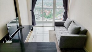 Un salon moderne avec un canapé gris, une table basse noire et un téléviseur posé sur un meuble. Une grande fenêtre offre une vue sur le paysage urbain.