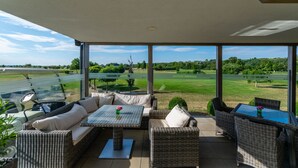 Eine überdachte Terrasse mit Korbmöbeln, einem Tisch mit einer Vase und Blick auf einen Golfplatz.