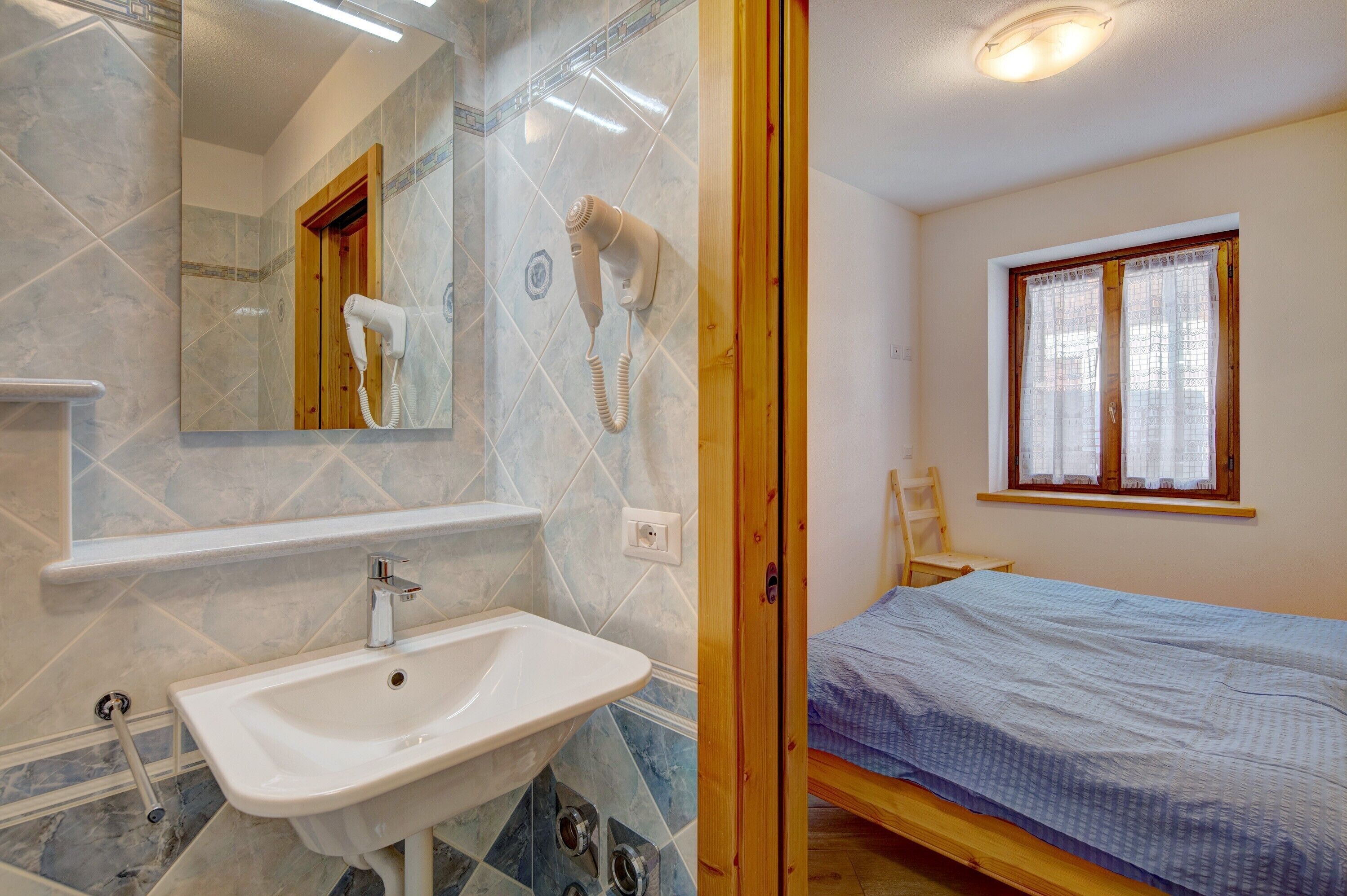 Ein Badezimmer mit Waschbecken, Spiegel und Haartrockner, ein Bett mit Fenster und eine Holztür.