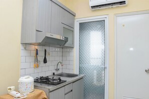 A compact kitchen with a gas stove, a microwave, and a sink. There is a small table with a kettle and a power strip. The walls are tiled, and there is a frosted glass door.