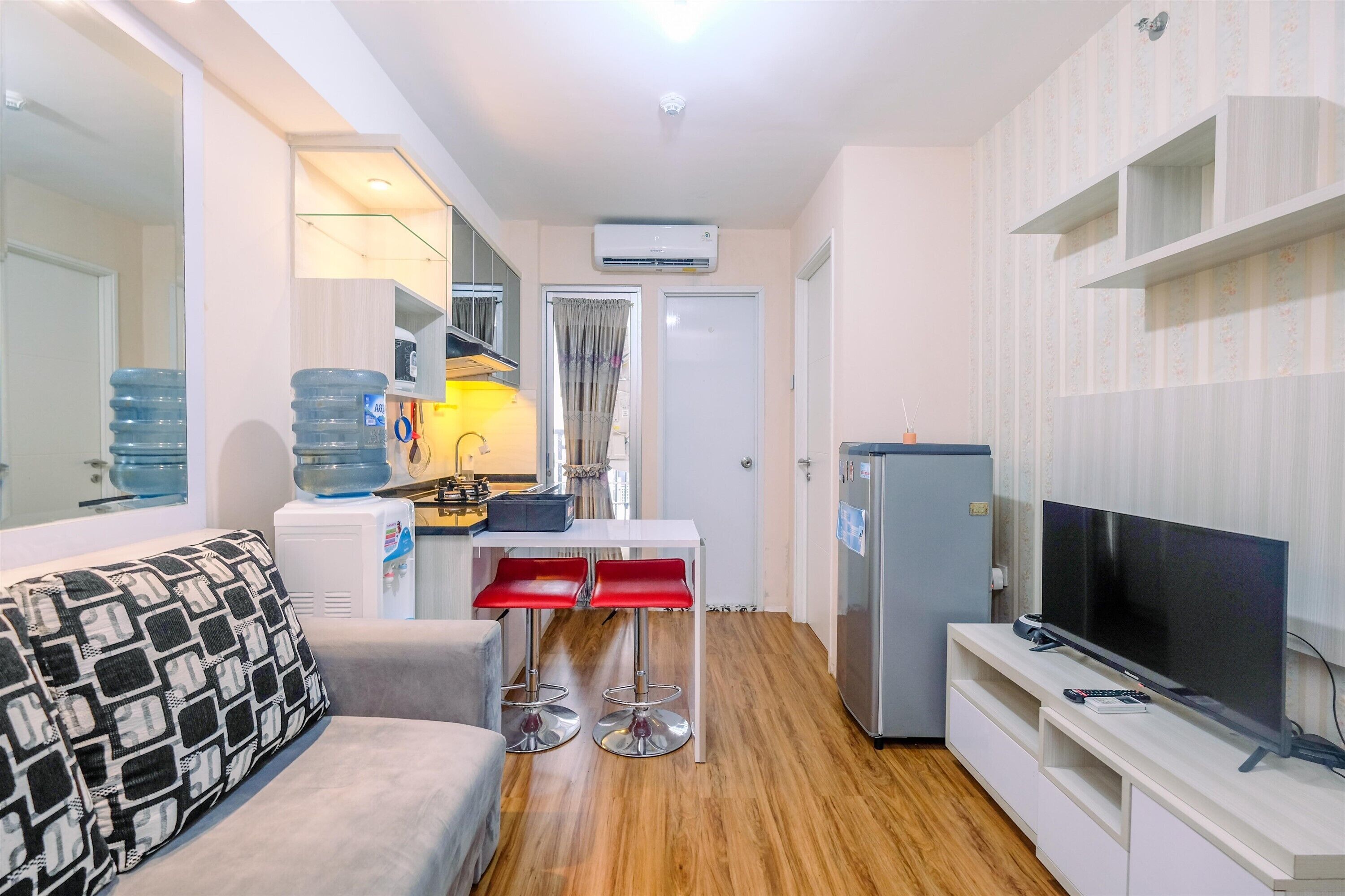 A compact living space with a sofa, a small dining table with red chairs, a TV on a white entertainment unit, and a kitchen area with a refrigerator and water dispenser.