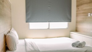 A bedroom with a bed, pillows, a bedside lamp, a window with a blind, and a wooden headboard. - Nice And Warm 1Br At Belmont Residence Puri Apartment (Jakarta)