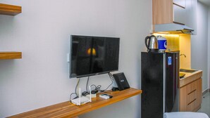 A compact hotel room with a flat-screen TV mounted on the wall, a small wooden desk with a computer and a coffee maker, a refrigerator with a water dispenser, and a wooden cabinet above the sink. - Nice And Cozy Studio At Evenciio Apartment (Depok)