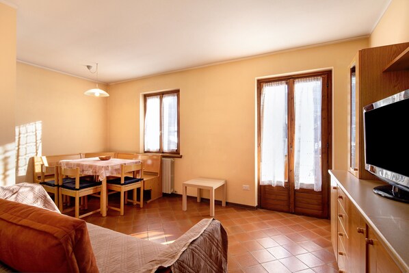 A room with a bed, a dining table with chairs, a small table, a television, and a window with curtains.