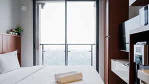 A hotel room with a large window, a bed with white linens, a wooden headboard, a TV on a wooden stand, and a small plant on the windowsill. - Cozy Studio Sky House Apartment Near Aeon & Ice Bsd (Pagedangan)