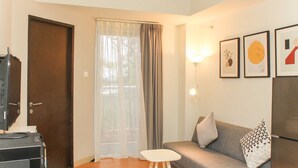 A living room with a grey sofa, a small white table, a door, and a window with curtains. - Nice And Elegant 1Br Apartment At Mustika Golf Residence (Cikarang)