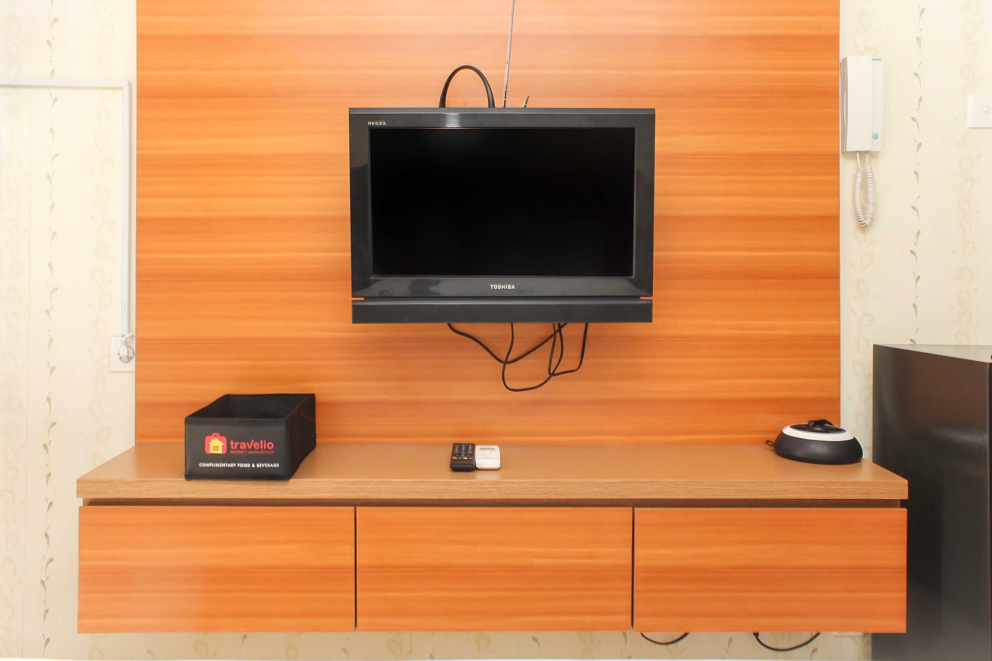 Un televisor de pantalla plana montado en una pared de madera, un minibar con una caja etiquetada 'Travelio', un control remoto y un altavoz sobre una encimera de madera.