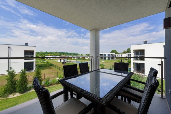 A balcony with a glass-top table and wicker chairs, overlooking a residential area with modern buildings and a body of water.