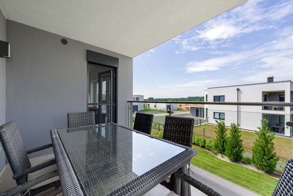Ein Balkon mit Glastisch und Korbstühlen, mit Blick auf einen modernen Gebäudekomplex und eine Rasenfläche.