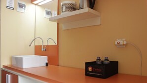 A bathroom with a white sink, two faucets, a mirror, a shelf with toiletries, and a traveler's box. - Cozy Studio Apartment At The Jarrdin Near Cihampelas Walk (Bandung)