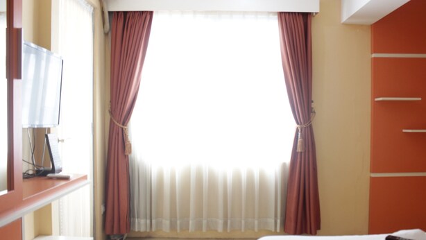 A hotel room with a bed, a TV on a stand, a window with curtains, and a wardrobe. - Cozy Studio Apartment At The Jarrdin Near Cihampelas Walk (Bandung)