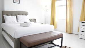 A modern hotel room with a large bed, a bedside table, a floor lamp, and a view of the outdoors through a window with yellow curtains. - Spacious And Modern Studio Room Apartment At Grand Asia Afrika (Bandung)