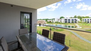 A balcony with a glass table and wicker chairs, overlooking a courtyard with a pond and modern buildings. - Apartment in Zsira near Thermal Spa (Zsira)