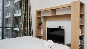 A hotel room with a bed, a television, a wooden shelf unit, and a window with curtains. - Elegant And Comfortable Studio Sky House Bsd Apartment (Pagedangan)