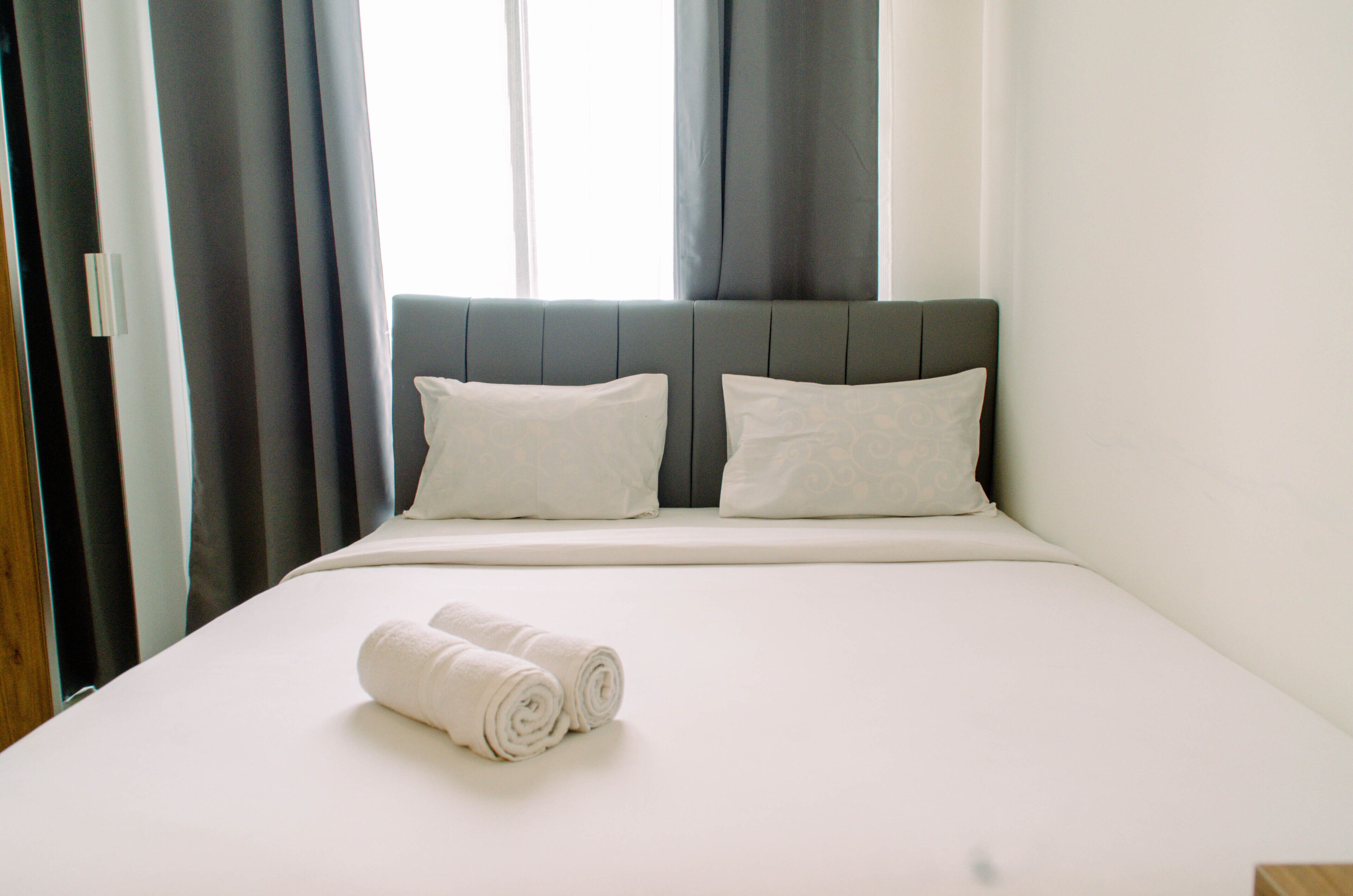 A neatly made bed with white linens and two pillows, a window with curtains, and a wooden door.