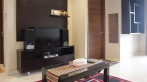 A living room with a flat-screen TV on a dark wooden stand, a small table with a striped table runner, and a door to the right. - Deluxe 2Br At Dago Boutique Apartment (Bandung)
