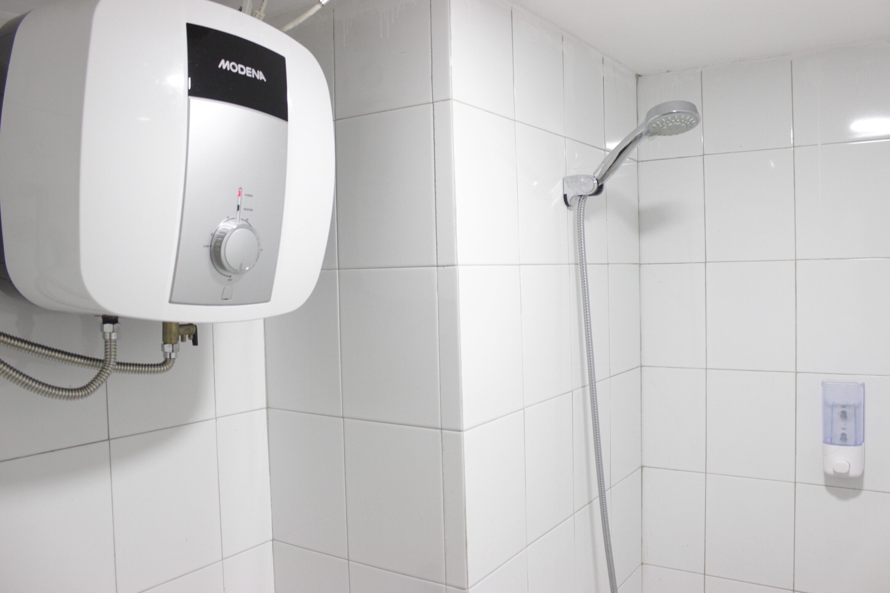 Ein weiß gefliester Duschbereich mit einer an der Wand montierten Duschbrause und einem weißen Warmwasserbereiter mit der Aufschrift 'MODENA'.