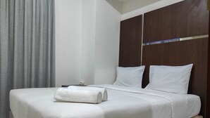 A neatly made bed with white linens and a rolled towel, a wooden headboard, and a grey curtain. - Chic And Cozy 2Br At Vida View Apartment (Makassar)